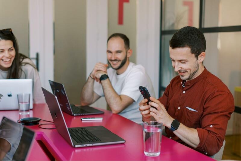 A Wolfpack Digital team member holding a smartphone at a pink desk beside an open laptop and a glass of water